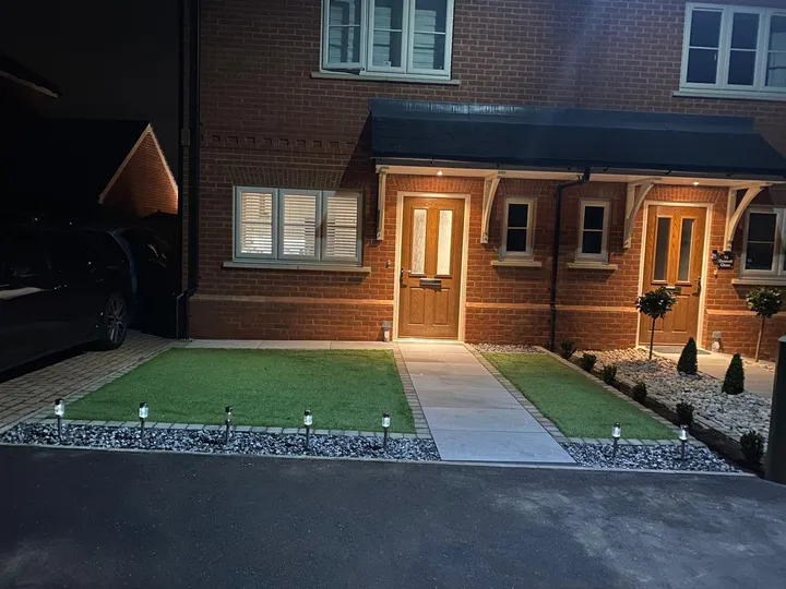 Front garden of a new-build lit at night — porcelain path, artificial lawn, clipped topiary and recessed ground lighting