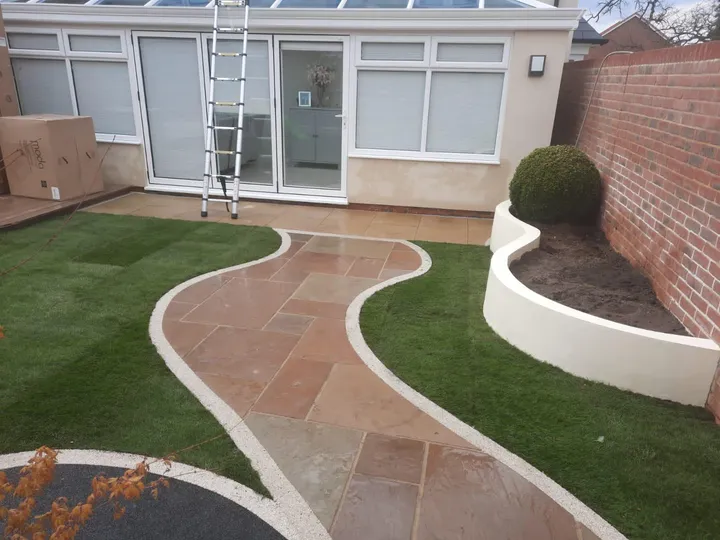 Sandstone path curving past a white rendered planter and circular bed to a conservatory