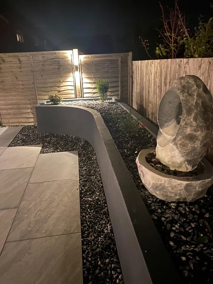 Carved stone water feature beside a rendered raised planter and slate mulch, lit at night by a wall-washing uplighter